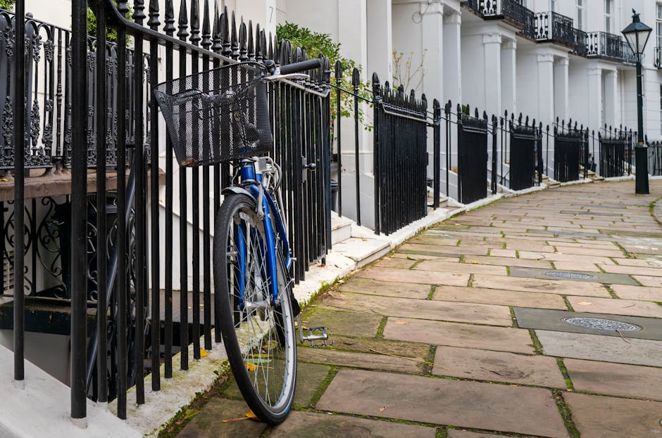 A paved sidewalk running alongside a row of elegant white terraced houses with black wrought iron fences. The fence is partially obstructed by a blue bicycle locked to it, with a front basket visible.
