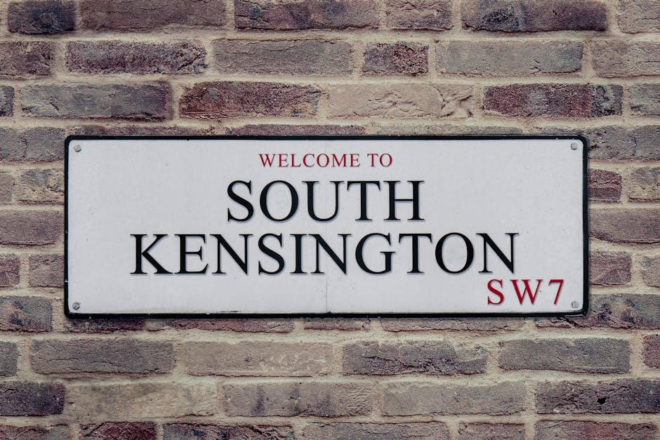 A rectangular street sign mounted on a red brick wall displaying the text 'Welcome to South Kensington SW7' in black and red lettering. The sign features a white background with black border edges, with the words 'South Kensington' prominently centered. The brick wall behind the sign has a rough texture with visible mortar joints between the bricks, and the lighting is natural, illuminating the sign and wall evenly. This image illustrates an outdoor scene typical of a residential or commercial area in South Kensington, with a focus on the signage that marks the location for visitors or residents. Cleaner South Kensington is familiar with maintaining such environments, ensuring cleanliness and proper signage for a well-kept neighborhood.