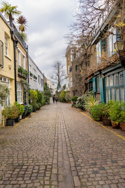 An outdoor cobblestone street in a residential area with row houses on each side, featuring a variety of exterior finishes including beige, white, and blue painted surfaces. The street is lined with potted plants and greenery, creating a lush and inviting atmosphere. Bare trees and overcast sky suggest a late autumn or winter season. In the distance, a few pedestrians are walking along the pathway. The scene is well-lit with natural daylight, highlighting the clean and well-maintained condition of the paved surface and surroundings. This image emphasizes the importance of surface cleaning and maintenance in urban residential environments, aligning with services offered by Cleaner South Kensington for domestic and commercial cleaning, as detailed in the Gloucester Road end of tenancy cleaning case study and costs.