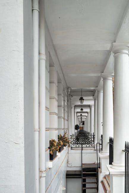 A row of white painted columns and walls forming a covered residential walkway or balcony with black metal railings and steps leading down. Potted plants with dried or wilted flowers are placed along