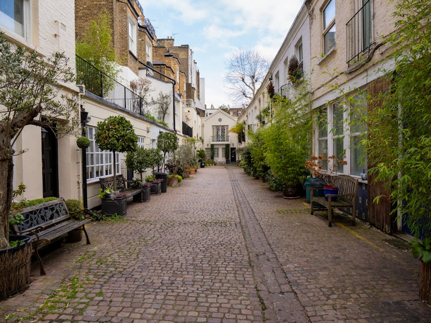 A narrow cobblestone courtyard flanked by white Victorian-style residential buildings with large windows and balconies decorated with potted plants and greenery. The courtyard features outdoor benches, and the ground appears clean and well-maintained. Some trees and shrubs in large planters add greenery to the space, while natural daylight illuminates the scene with a clear sky overhead. The area exudes a tidy, inviting residential atmosphere, typical of South Kensington, and reflects the kind of surface cleaning and maintenance highlighted in the Kensington High Street carpet cleaning guide for homeowners, provided by Cleaner South Kensington.