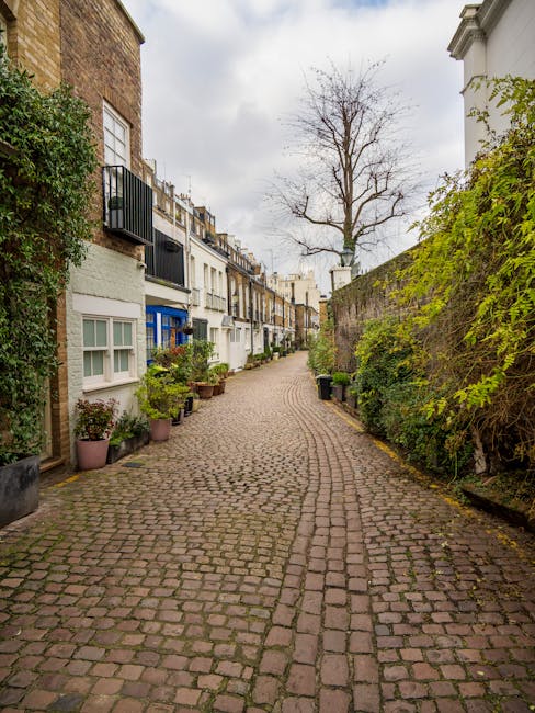 A narrow cobblestone street in South Kensington, lined with residential houses featuring white and colored facades, some with small balconies and window boxes. Potted plants and greenery are arranged along the sidewalk, with a leafless tree standing in the distance under an overcast sky. The natural lighting highlights the textured stone surface and clean appearance of the street, exemplifying a well-maintained area. This scene reflects the typical charming and tidy atmosphere of South Kensington, suitable for highlighting professional cleaning services by Cleaner South Kensington for Airbnb hosts. No visible cleaning tools or surfaces are present in the image.