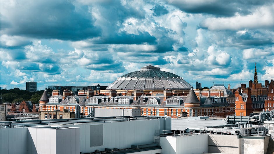 View of the rooftop of a historic venue, including a large circular glass and metal dome structure surrounded by Victorian brick buildings with turrets and gabled windows, under a partly cloudy sky. The rooftop surfaces are finished with white and gray metal panels, reflecting sunlight. This setting illustrates the importance of diligent surface cleaning and maintenance for event spaces like the Royal Albert Hall. Cleaner South Kensington provides professional cleaning services, ensuring high standards of hygiene, surface shine, and dust removal for venues of this scale. The image emphasizes the need for regular deep cleaning and sanitisation of both exterior and interior surfaces to maintain a pristine appearance for public events.