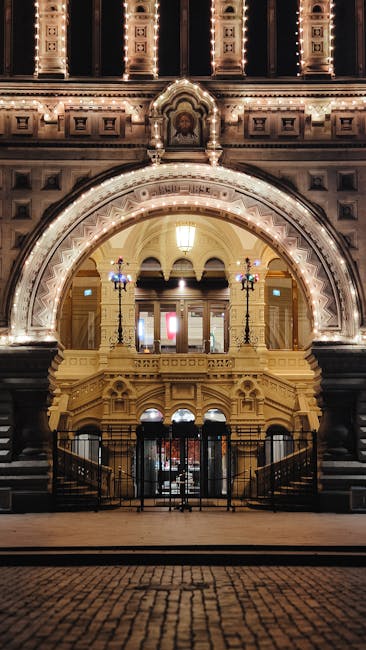 The image shows the ornate entrance of the Royal Albert Hall, featuring a large arched doorway decorated with detailed architectural moldings and illuminated by warm lighting. The facade includes decorative stonework, columns, and intricate trim, all in a classical style. Two vintage-style street lamps flank the entrance, casting a soft glow on the steps below. The area appears clean and well-maintained, with polished surfaces and no visible dust or debris. The lighting highlights the texture of the stonework and the elegant design of the venue, providing an inviting and majestic appearance. This setting exemplifies the importance of surface cleaning and maintenance in preserving the grandeur of historic venues, aligning with the focus of professional cleaning services by Cleaner South Kensington for event spaces like the Royal Albert Hall.