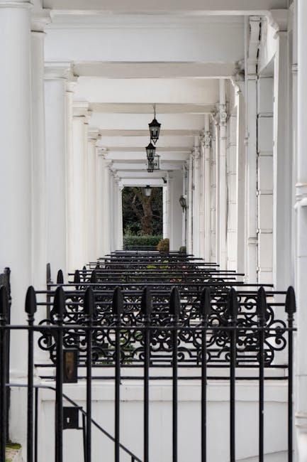 A row of white painted wooden columns and beams forming a covered corridor with a black decorative wrought iron fence in the foreground. The corridor features classic pendant lanterns hanging from the ceiling, and the flooring appears clean with a smooth surface. In the background, a view of greenery can be seen through the open end of the corridor. The overall scene emphasizes a well-maintained, hygienic outdoor space, indicative of thorough surface cleaning and maintenance, as provided by Cleaner South Kensington, to ensure cleanliness and presentation for residential or commercial exteriors.
