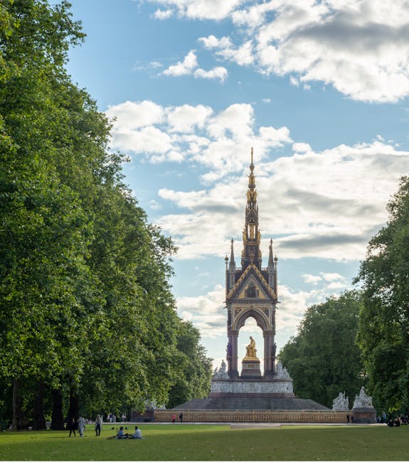 An outdoor scene in a park showing a historic monument with a tall, ornate spire and a golden statue at the base, surrounded by lush green trees. The monument features intricate gothic architecture with decorative elements and is positioned on a stone platform. The sky above is partly cloudy with patches of blue, and a few visitors are seen relaxing on the grass in the foreground. This image highlights natural stone and metal surfaces, clean and well-maintained, with sunlight illuminating the monument and greenery. As part of [COMPANY_NAME]'s cleaning offerings, such detailed surface cleaning and maintenance ensure these historic structures are preserved and presented in a pristine condition, aligning with the focus of the South Kensington cleaning cost guide for SW7 residents.